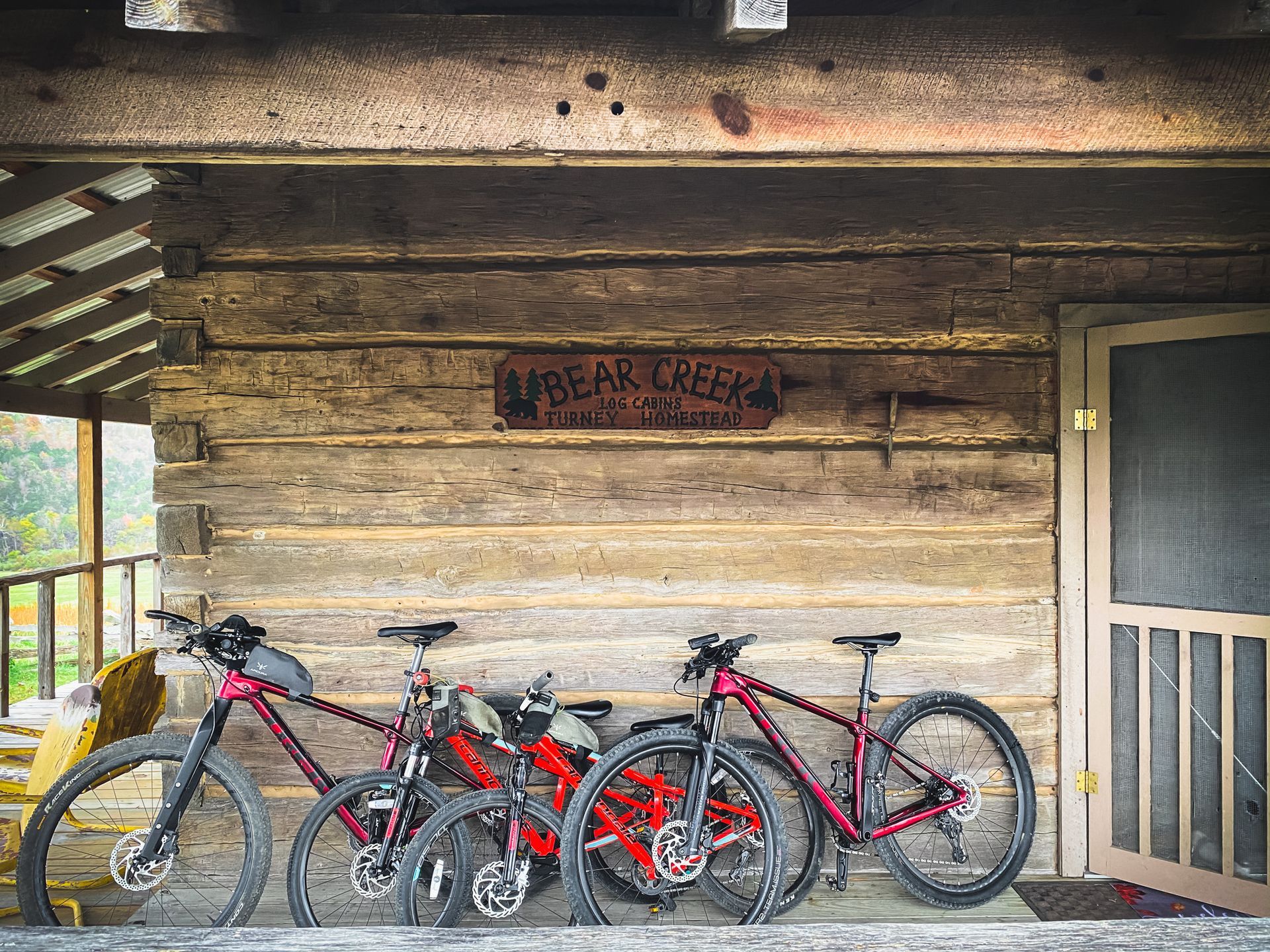 Three mountain bikes parked under the porch of a log cabin with a