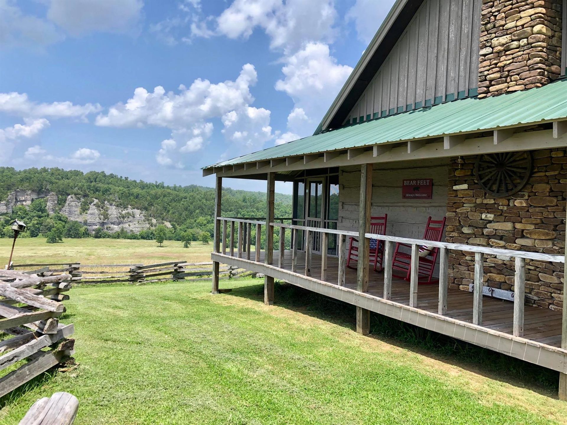 Cabin with green roof and stone chimney overlooking a grassy field and distant cliffs.