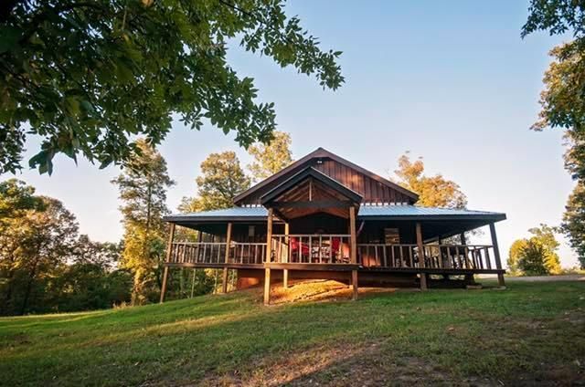 Wooden cabin with wraparound porch on a grassy hill, surrounded by trees.
