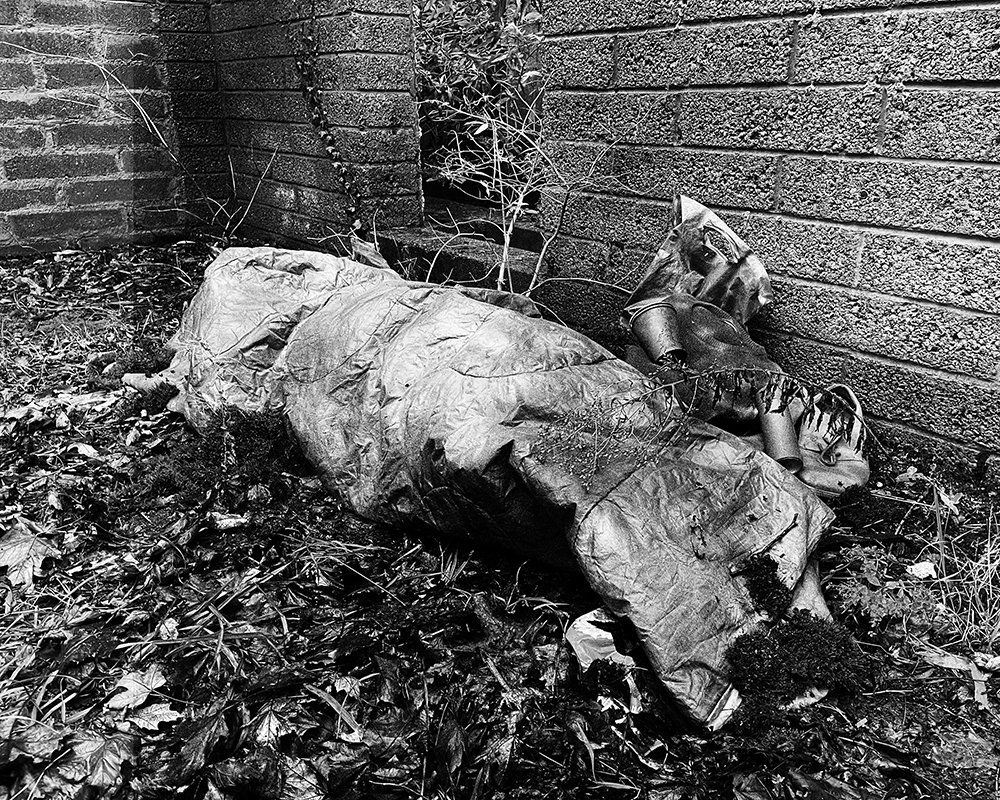 An empty sleeping bag, carefully posed to resemble a resting body, lies in a forgotten alley corner.