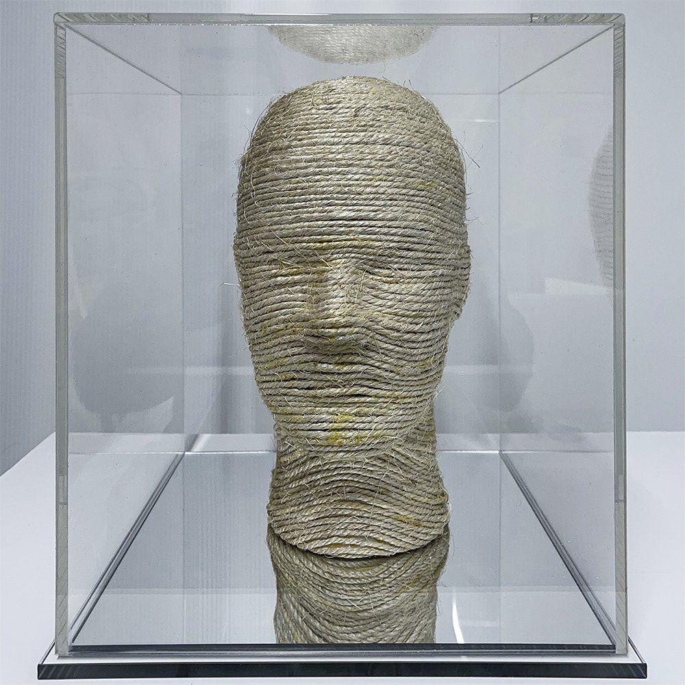 A discarded mannequin head wrapped in sisal, enclosed in a clear acrylic box with a mirrored base reflecting the viewer.