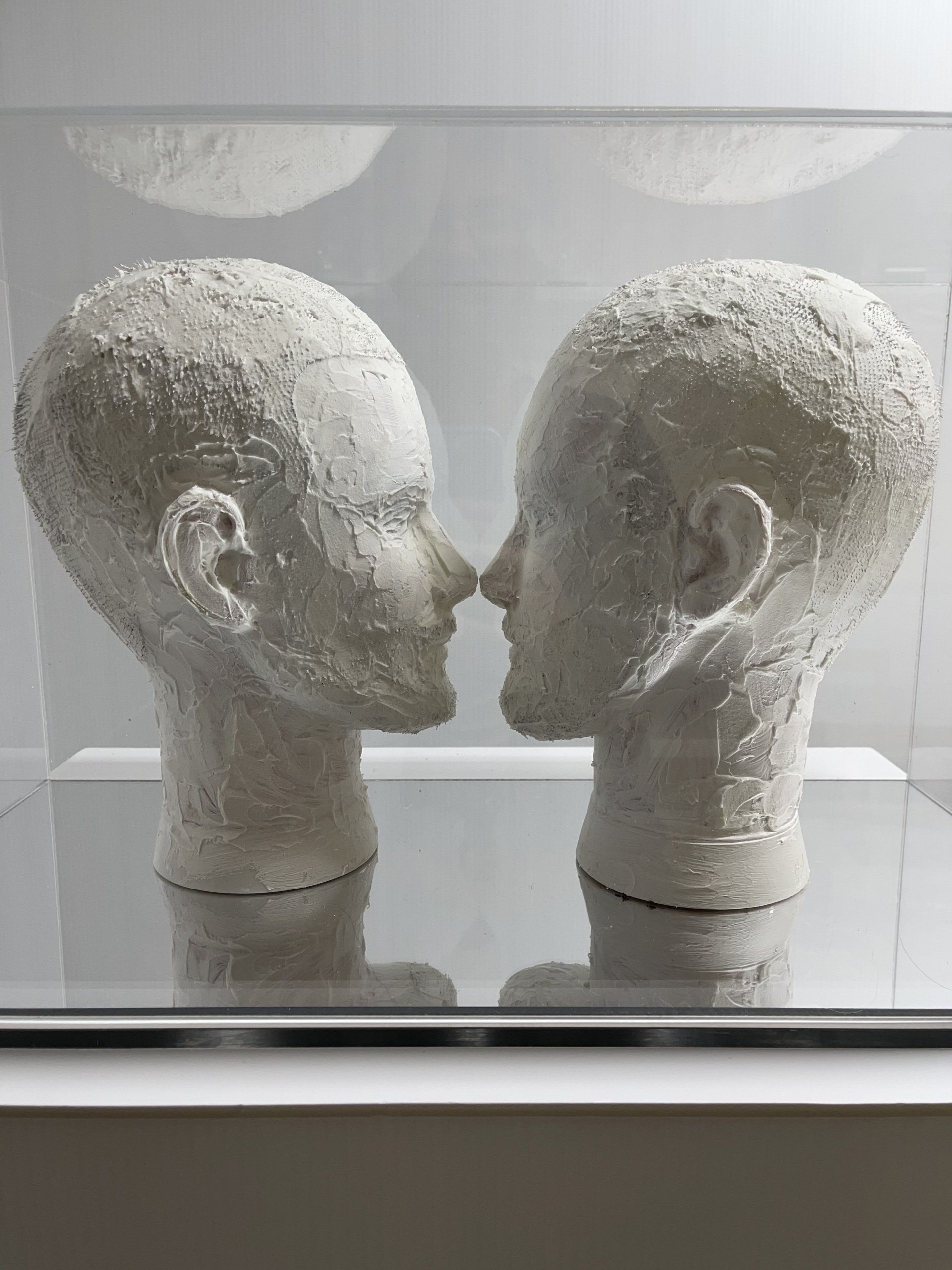 Two plaster covered mannequin headspace each other, enclosed in a clear acrylic box with a mirrored base reflecting the viewer.