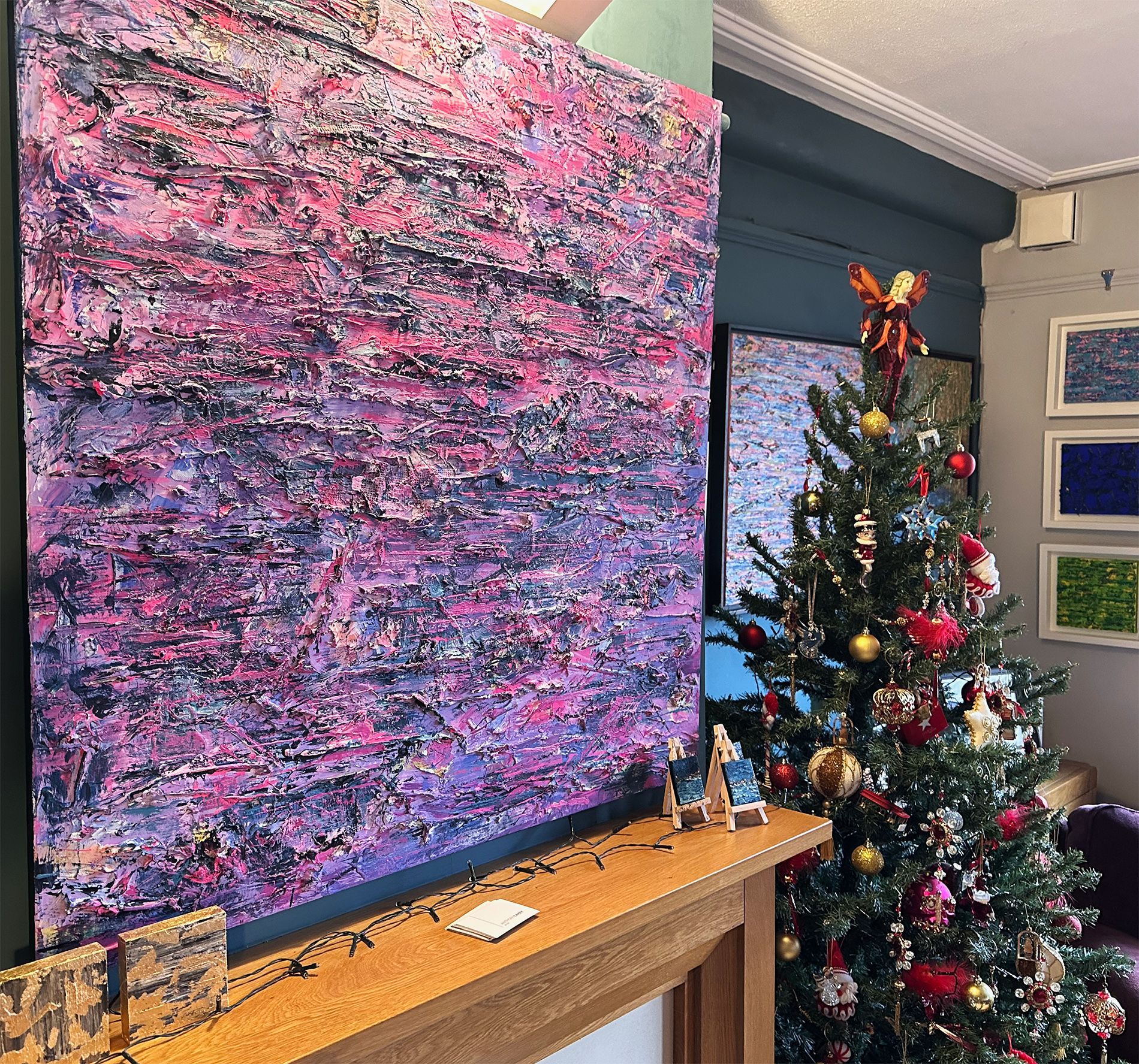 Extra large abstract painting in deep pinks and blues hanging above a fireplace.There is a decorated Christmas tree in the background and some smaller paintings visible