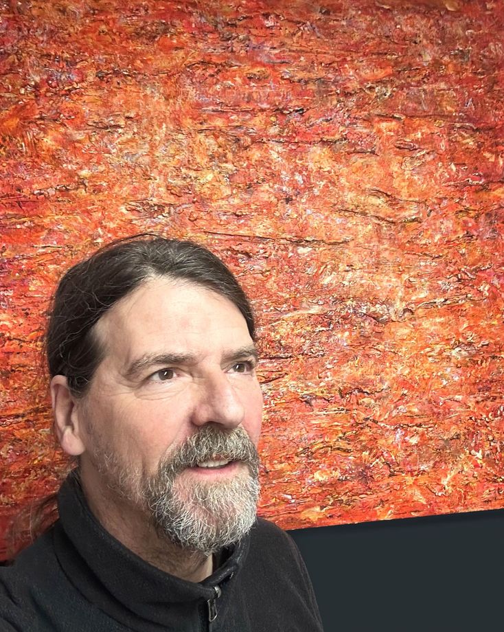 A white bearded male, the artist Anthony Carey standing in front of one of his latest works. A large richly textured canvas in  deep oranges and yellows