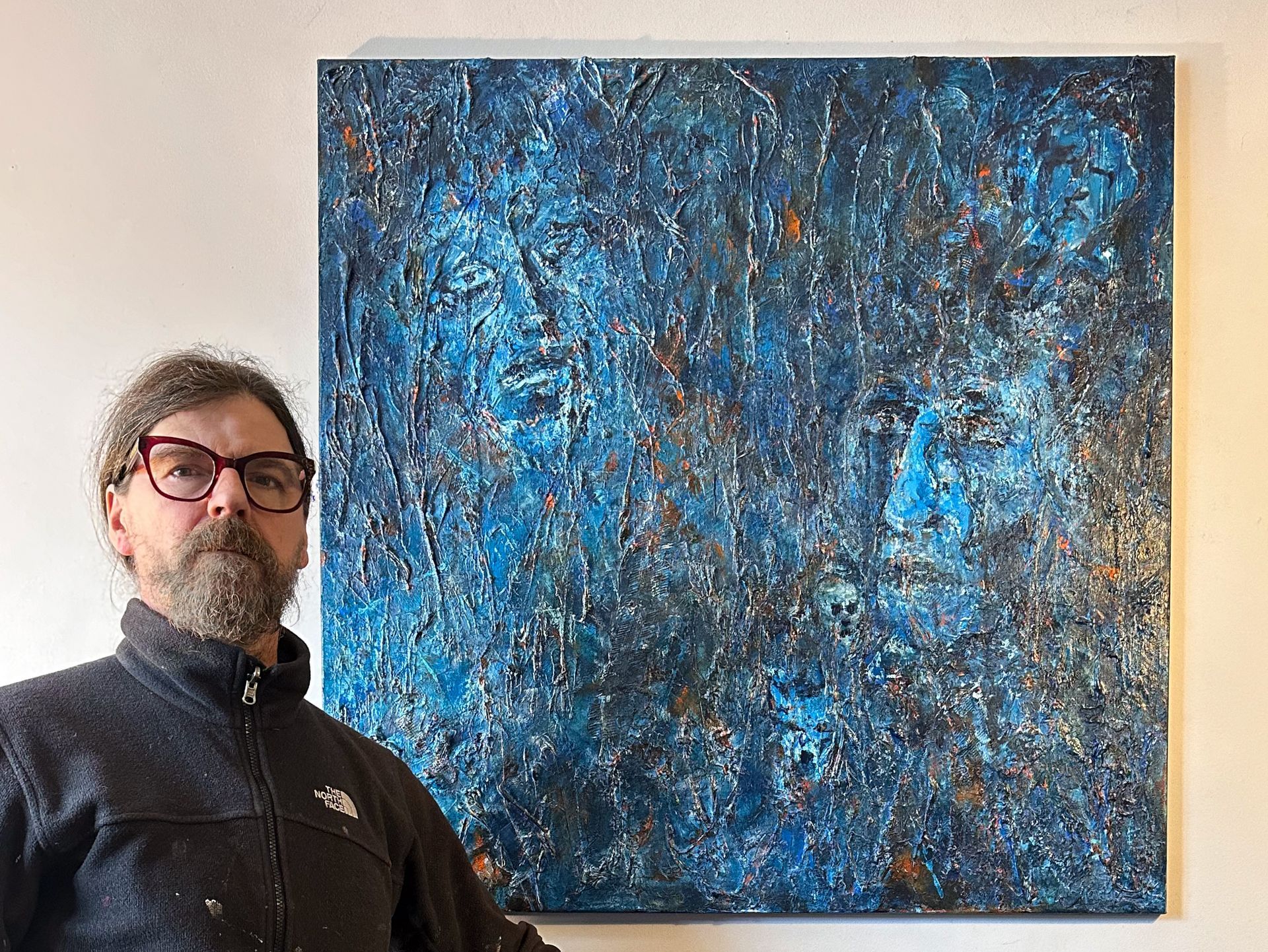 The artist Anthony Carey a bearded white male beside his latest painting, 'The Ancestors', a large painting in dark blue showing two faces emerging from the shadows 