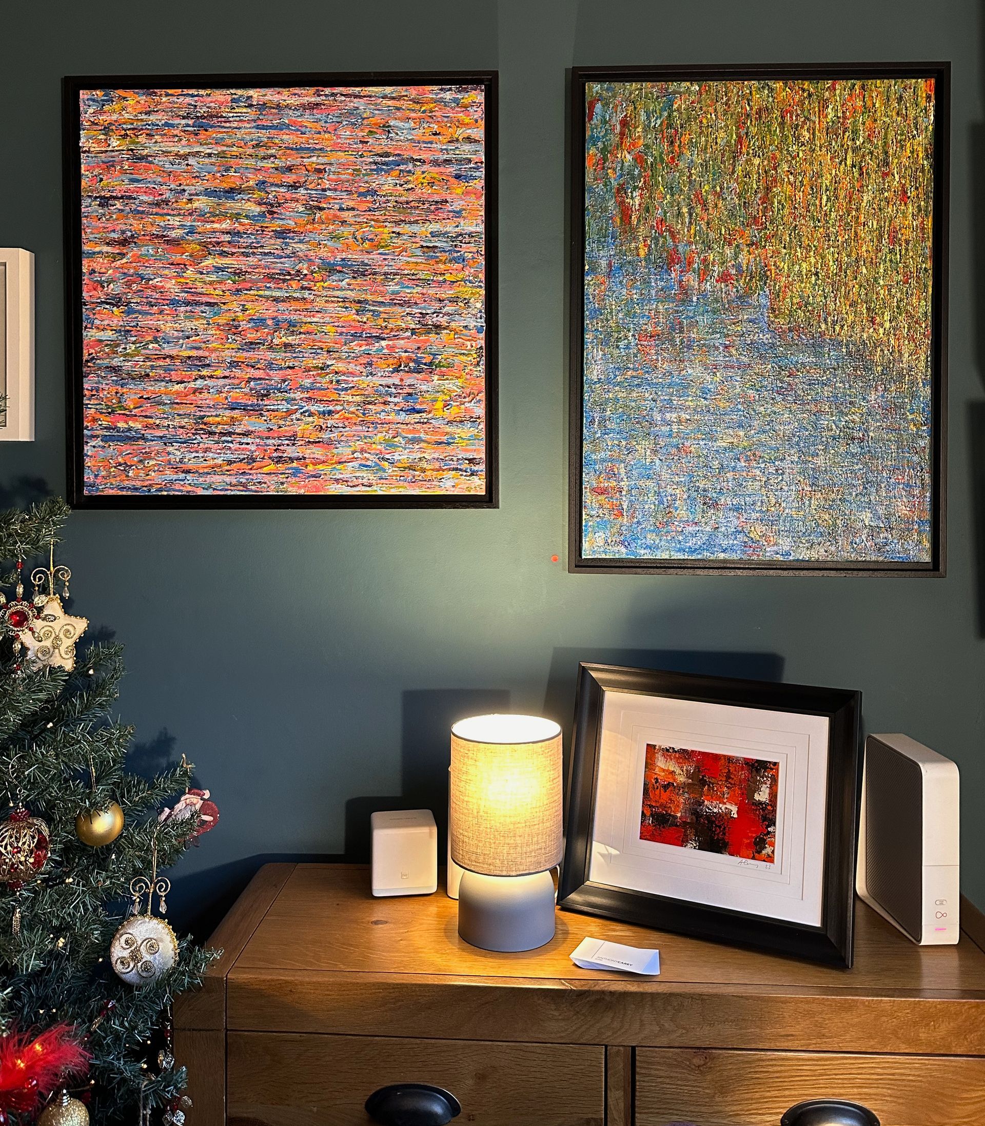Two large abstract paintings hang on the artists wall. in the foreground is a side table with a small lamp and a framed small abstract painting in a black frame