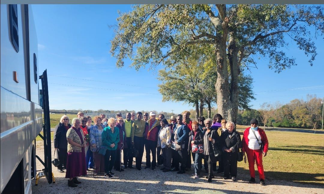 Group of people posing by a large tree on a sunny day next to a parked bus.