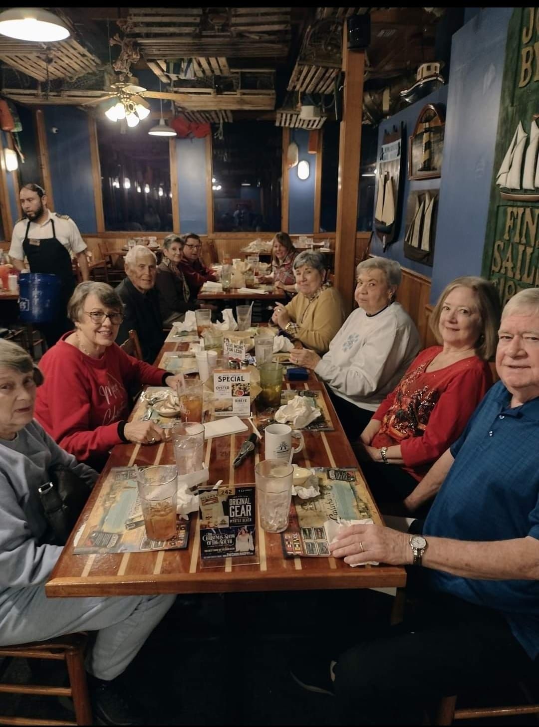 Group of people seated around a table in a restaurant. Server nearby. Wooden table tops, blue walls.
