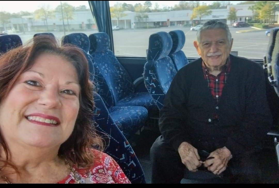 Woman takes a selfie with an older man on a bus. They are smiling. Bus interior and parking lot visible.