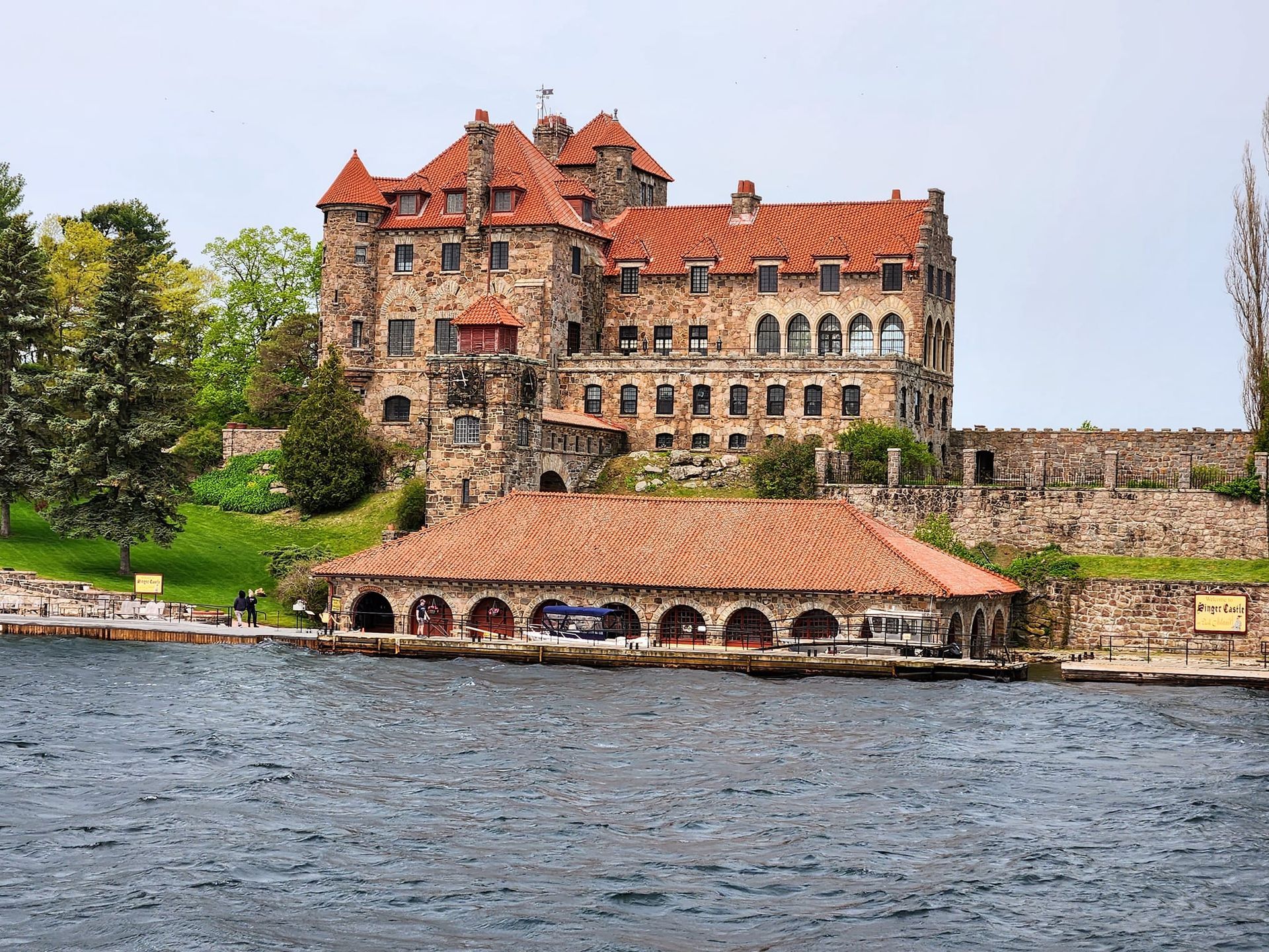Stone castle with red-tiled roof and boathouse by water. Overcast day.