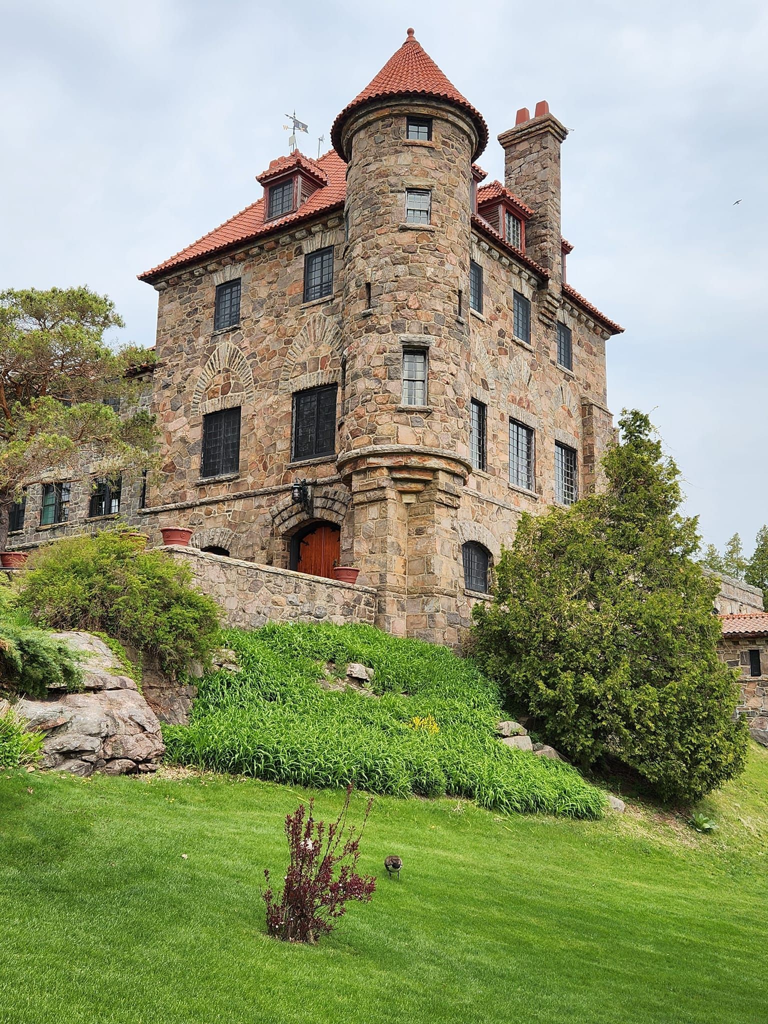 Stone castle with red tile roof and round turrets on a green hillside with landscaping.