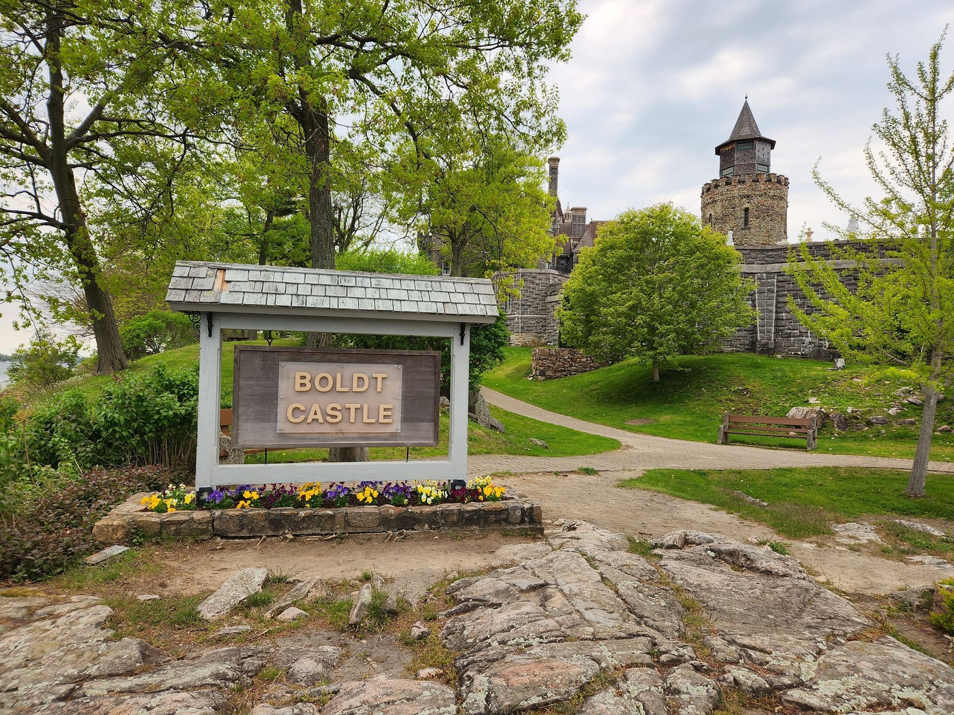 Sign for Boldt Castle, with stone castle in background, trees and cloudy sky.