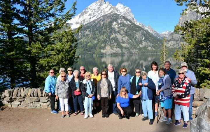 Group of people pose by a lake with a snow-capped mountain backdrop. Bright sunny day.