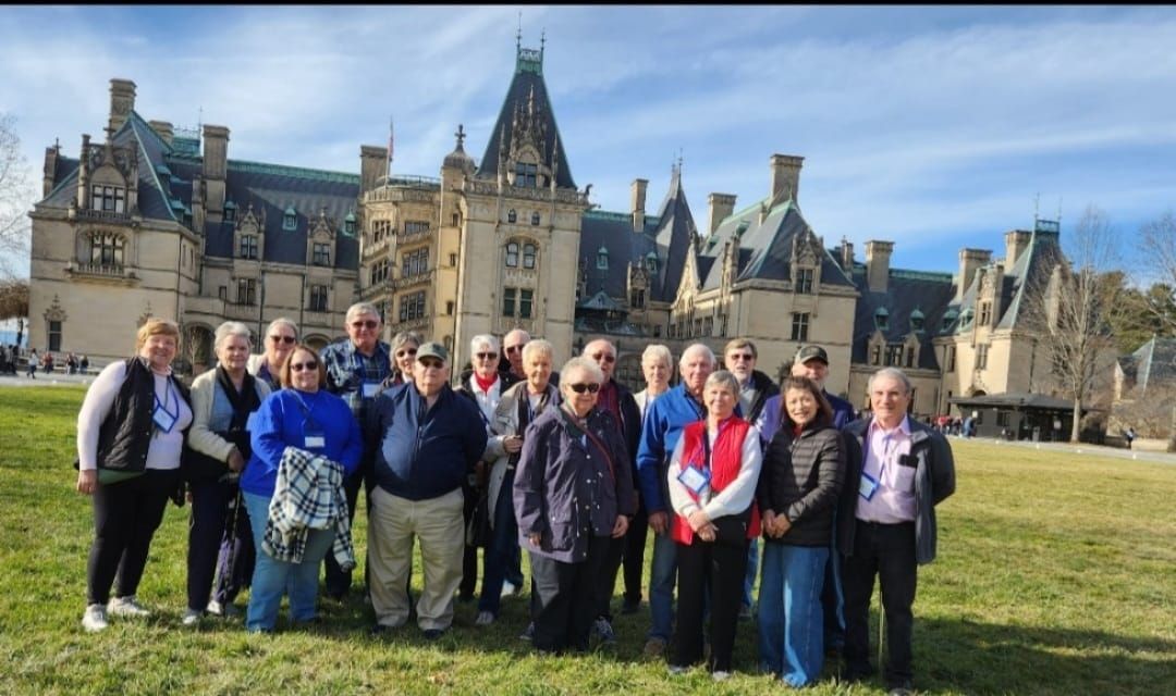 Group of people posing in front of the Biltmore Estate, a large castle-like building. Sunny, outdoor setting.
