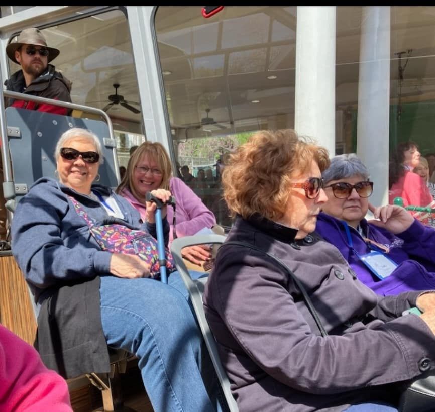People seated on a tour bus, including a woman with a cane, some wearing sunglasses, with a man in the background.