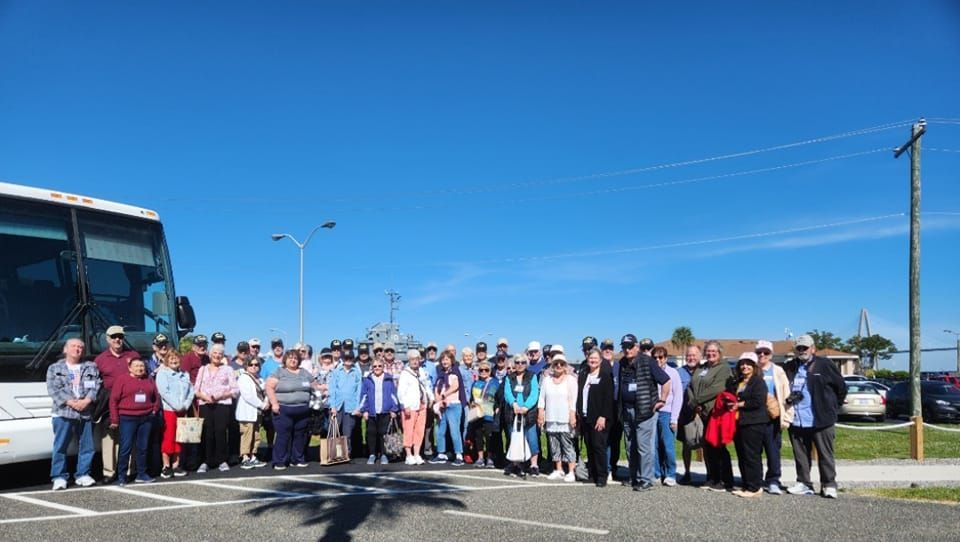Group of people standing in front of a bus on a sunny day. They are wearing casual clothes and some have lanyards.