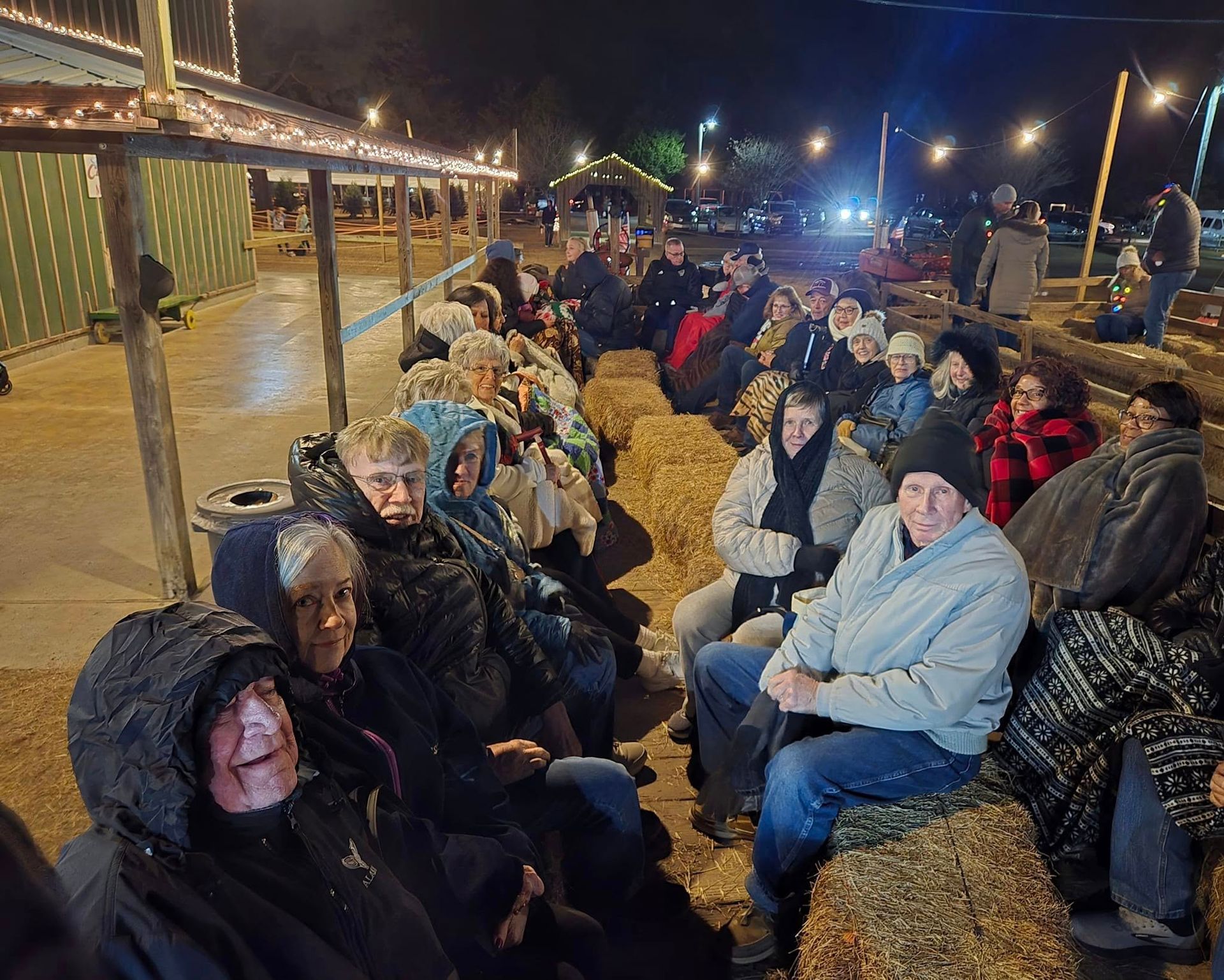 People sit on hay bales, bundled in coats, under string lights. Outdoor night scene.