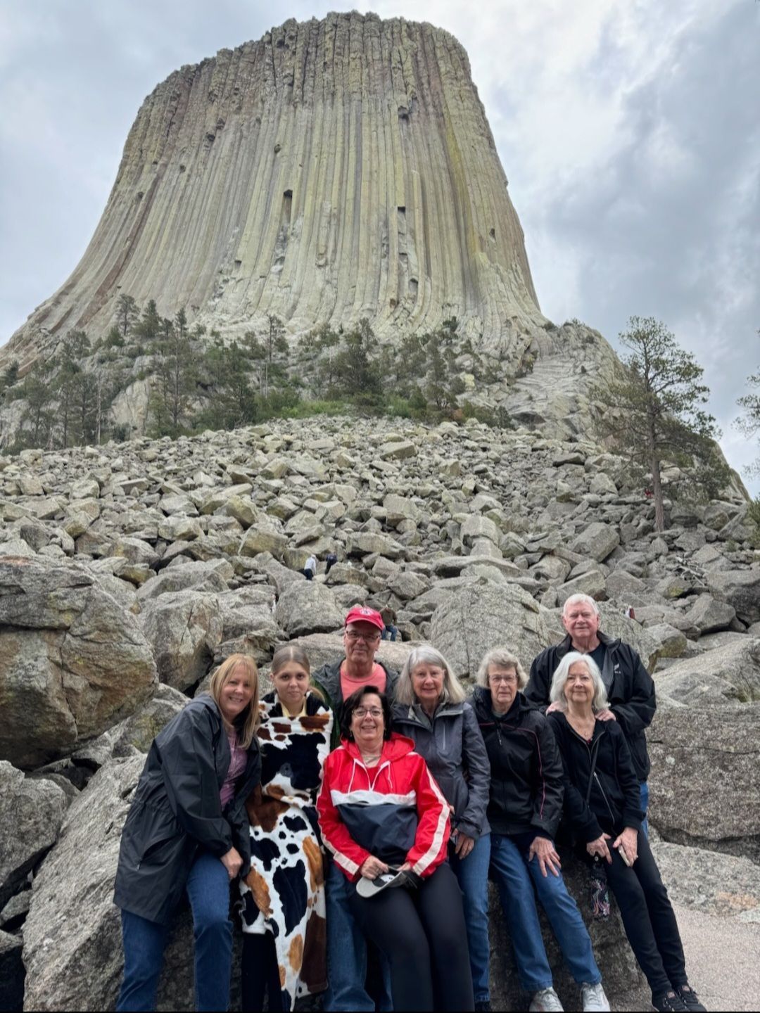 Group of people posing in front of Devils Tower, Wyoming, a large rock formation under a cloudy sky.