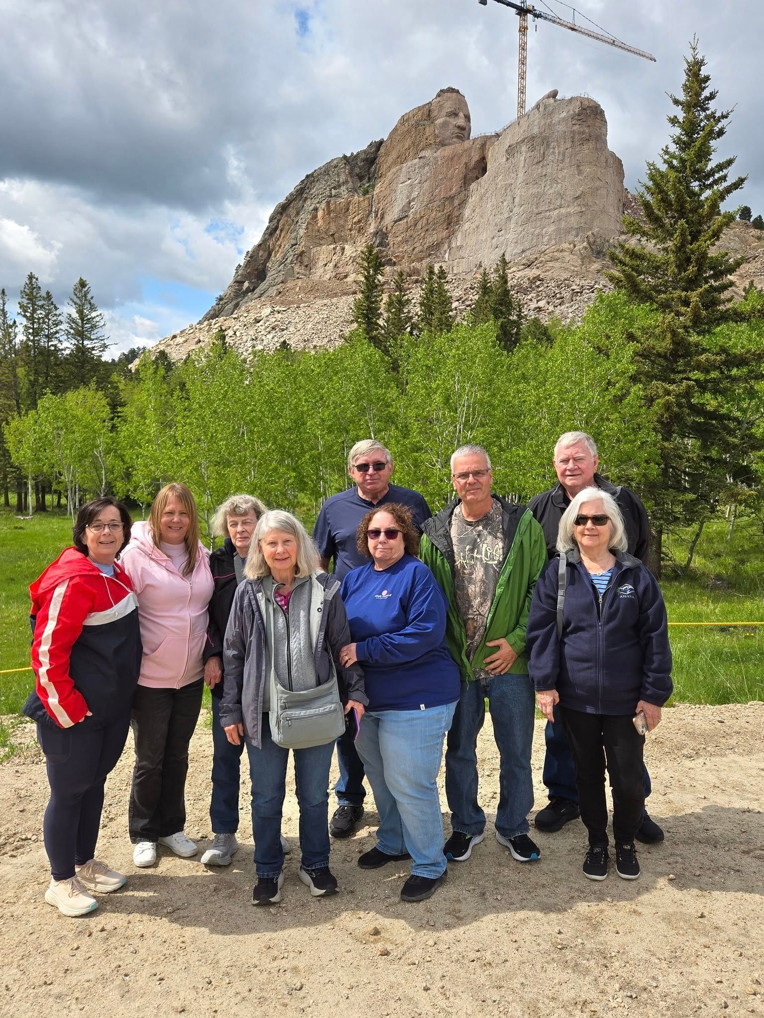 Group of people pose in front of a mountain with a crane at the top. Green trees, cloudy sky.