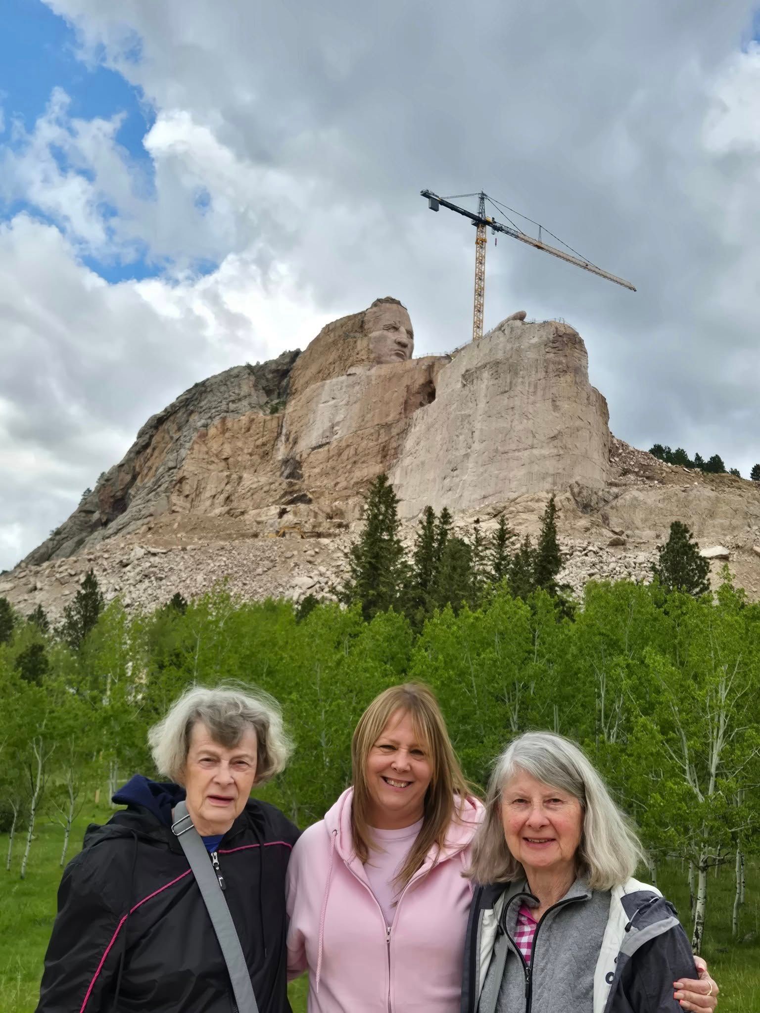 Three people pose in front of the Crazy Horse Memorial, a mountain carving in South Dakota.