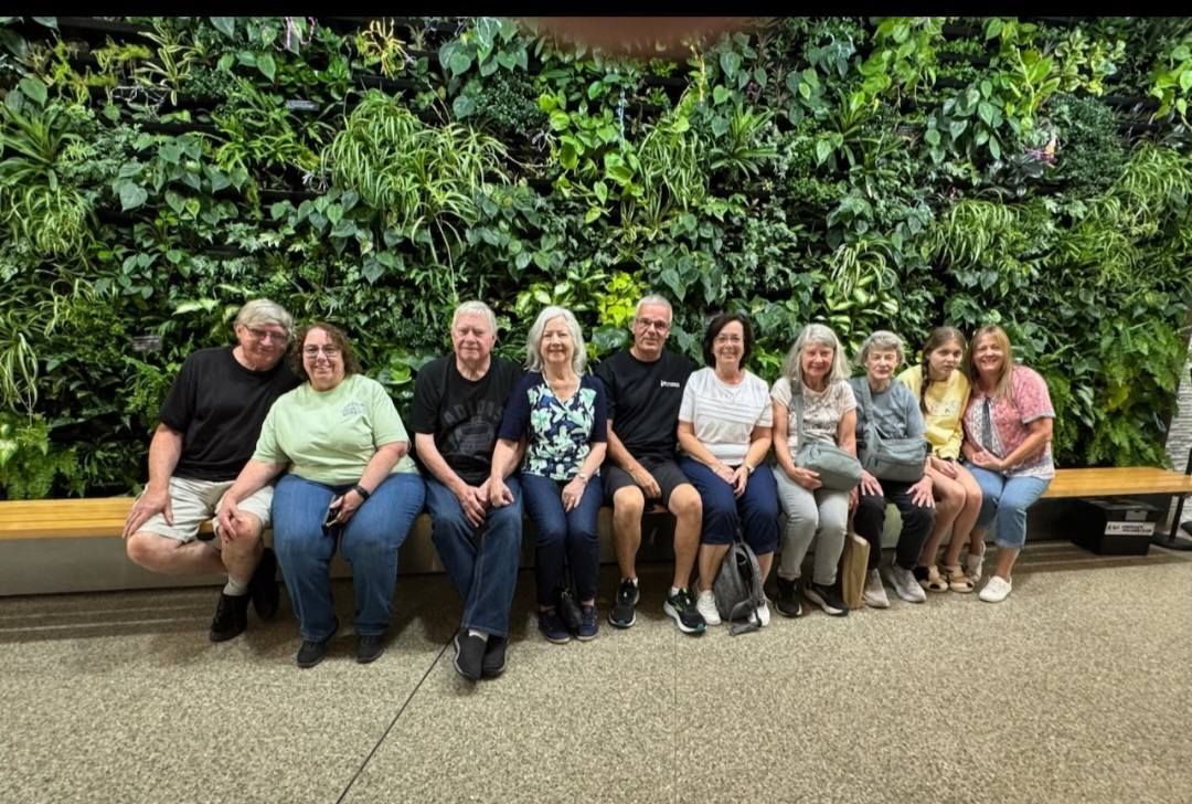 Group of people sitting on a bench in front of a green wall of plants.