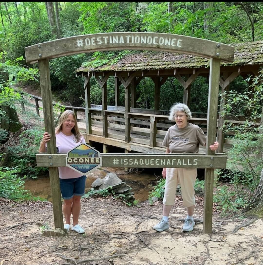 Two people pose in a wooden frame with a bridge and greenery. The frame reads #DESTINATIONOCONEE and #ISSAQUEENAFALLS.