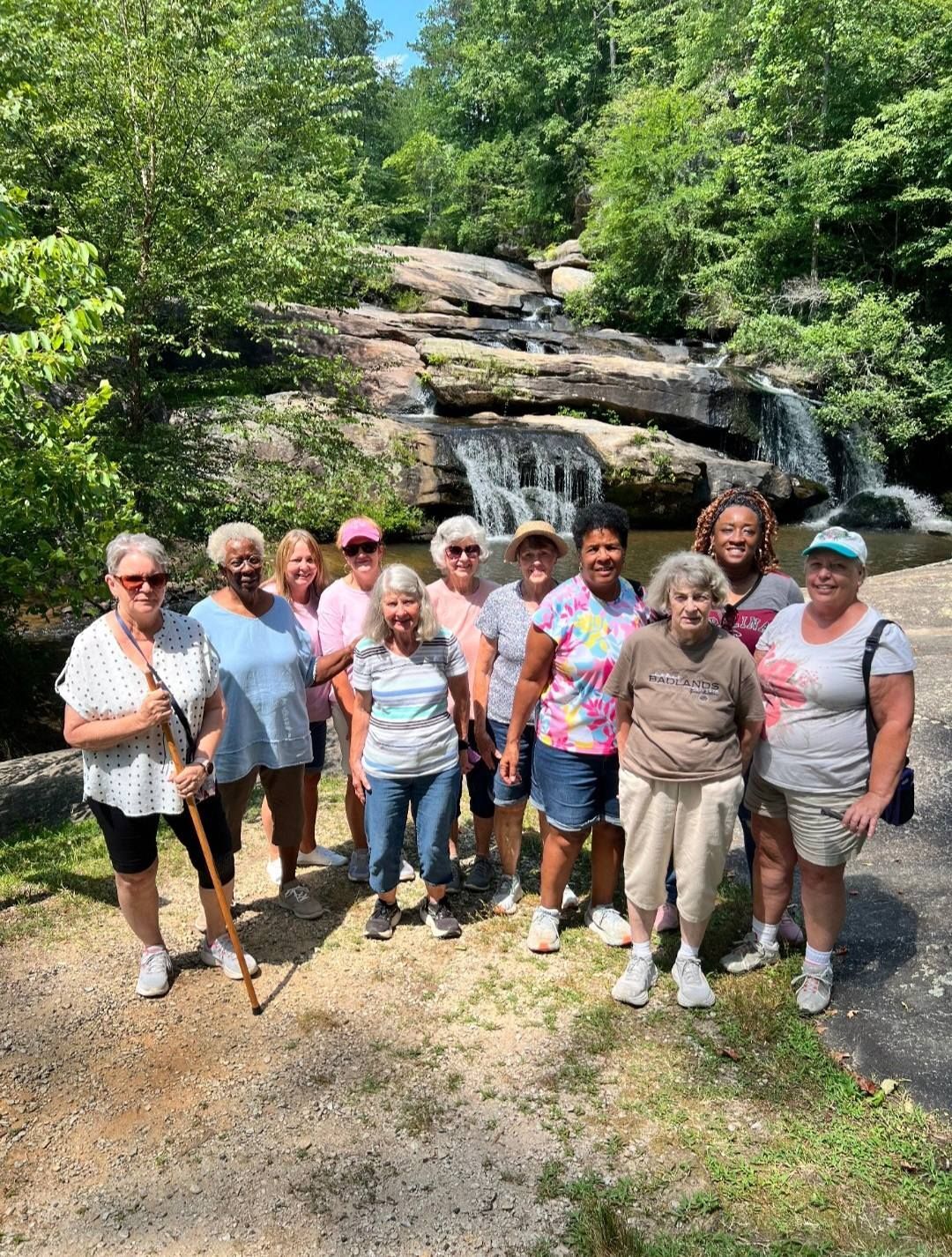 Group of people standing near a small waterfall, sunny outdoor setting.