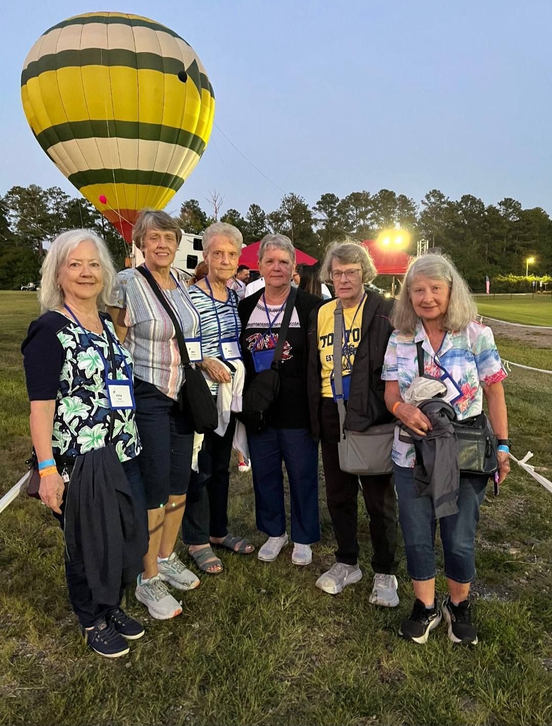 Group of women pose in front of a yellow and white hot air balloon on a grassy field at dusk.
