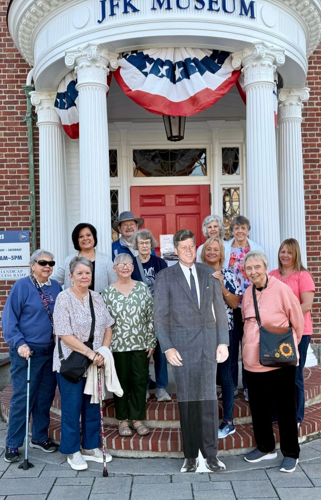 Group of people posing with a JFK cutout in front of the JFK Museum, decorated with flags.