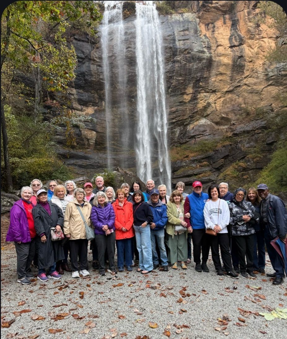 Group of people standing in front of a tall waterfall with a rocky cliff backdrop.