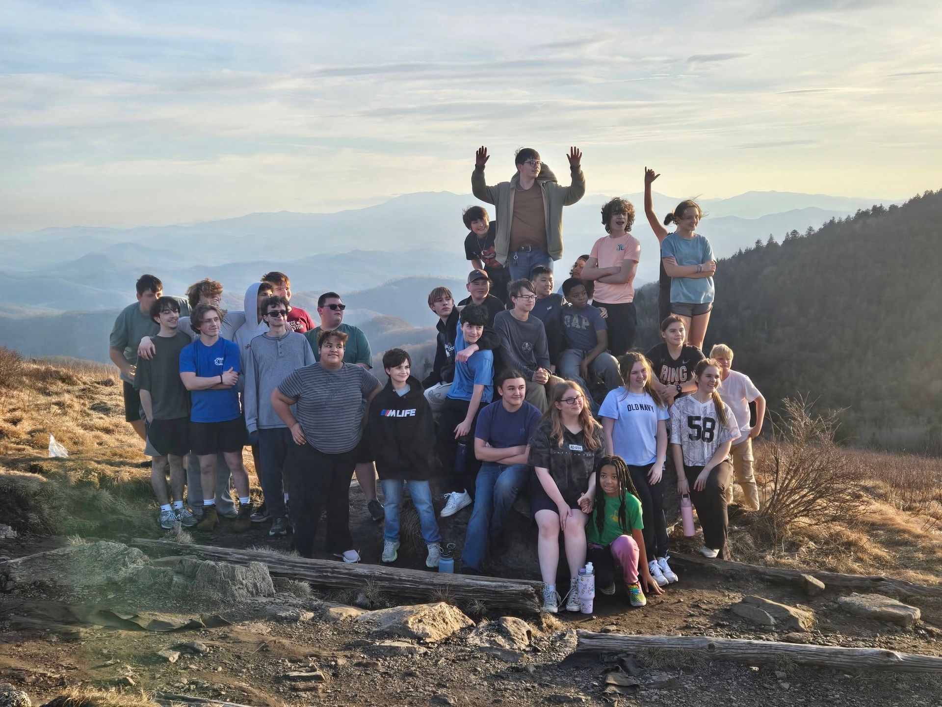 Group of people on mountaintop, arms raised, posing. Blue and gray hues, mountainous backdrop.
