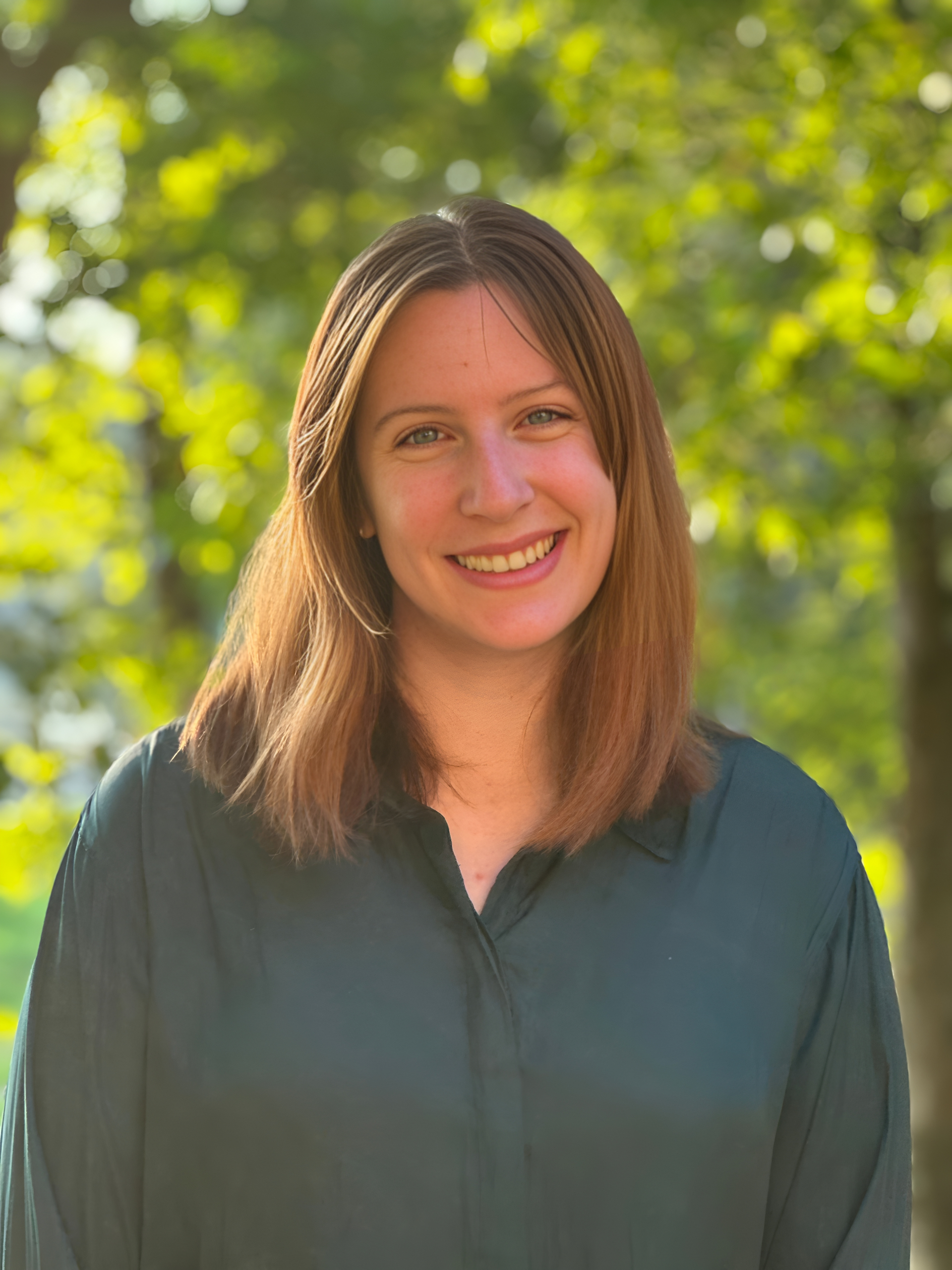 A woman in a green shirt is smiling for the camera in front of trees.