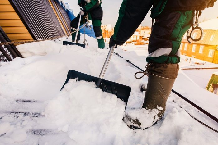 Person shoveling snow off a rooftop, wearing boots and safety gear.