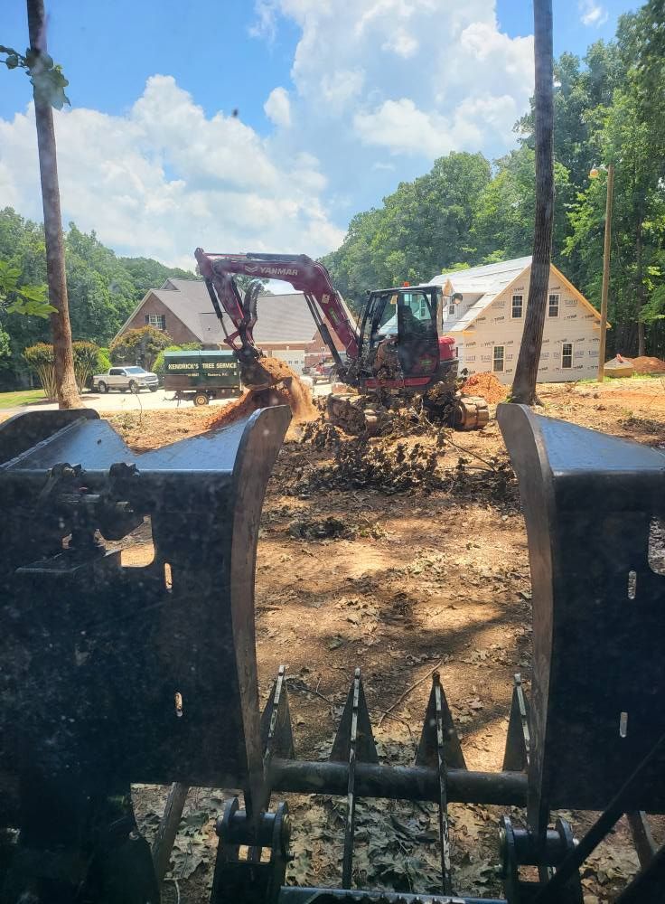 Excavator digging dirt at a construction site with a partially built house and trees in the background under a blue sky.