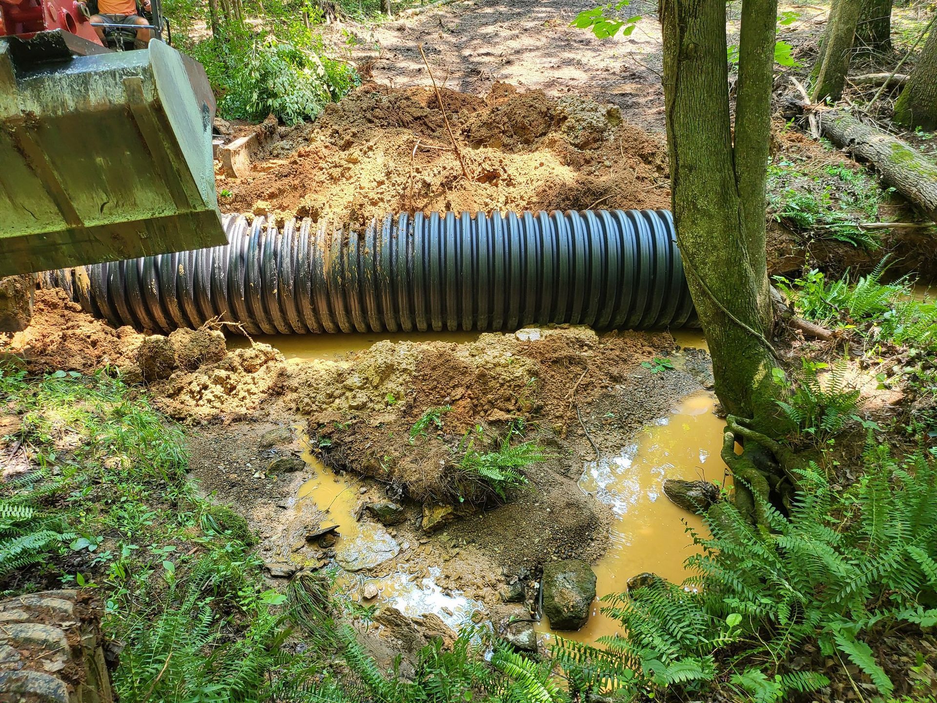 Excavator installing a large corrugated pipe in muddy forest stream.