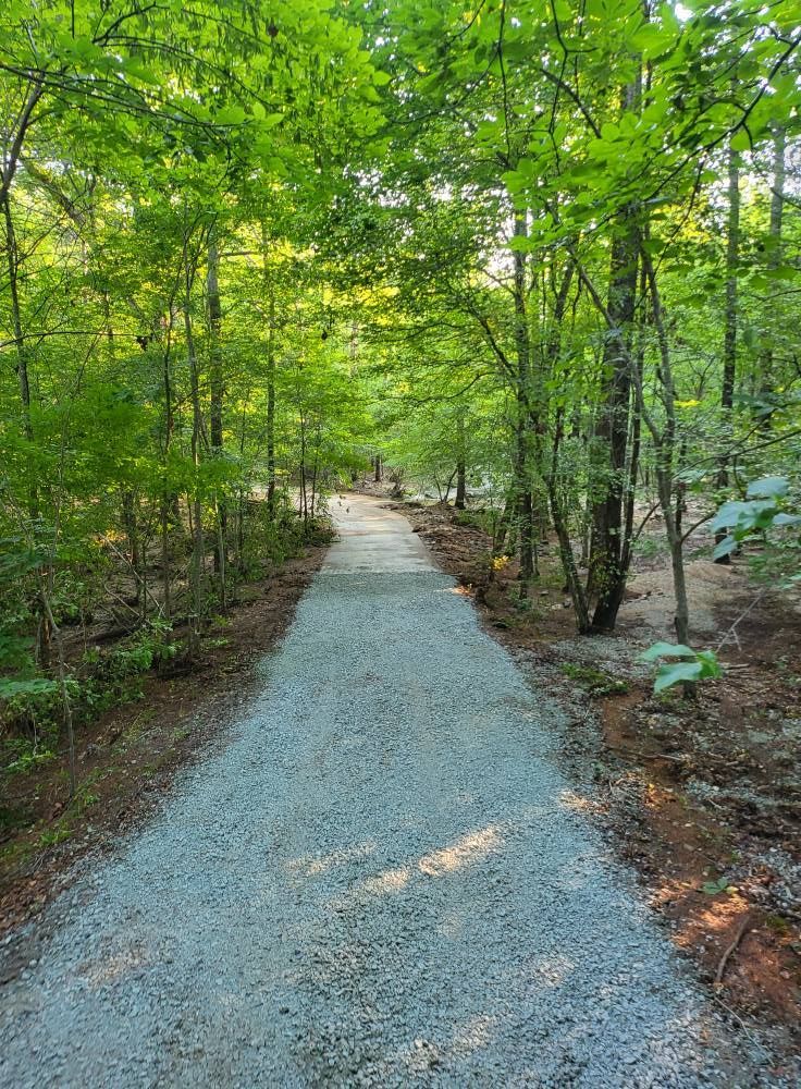 Gravel path winding through a forest. Trees with lush green leaves line the trail.