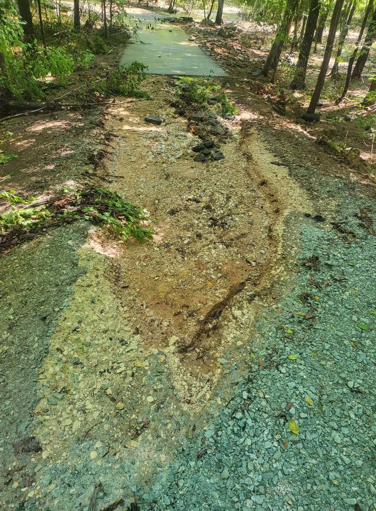 Erosion damage on a gravel path in a wooded area, leading to a concrete surface.
