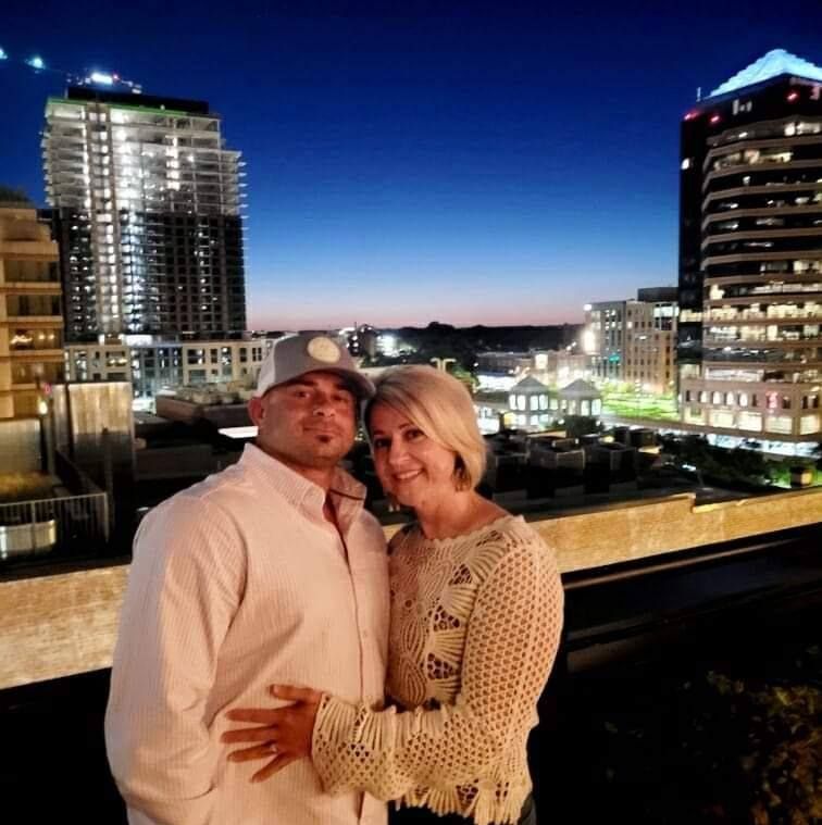 Couple hugging, posing on rooftop with city skyline backdrop at dusk.