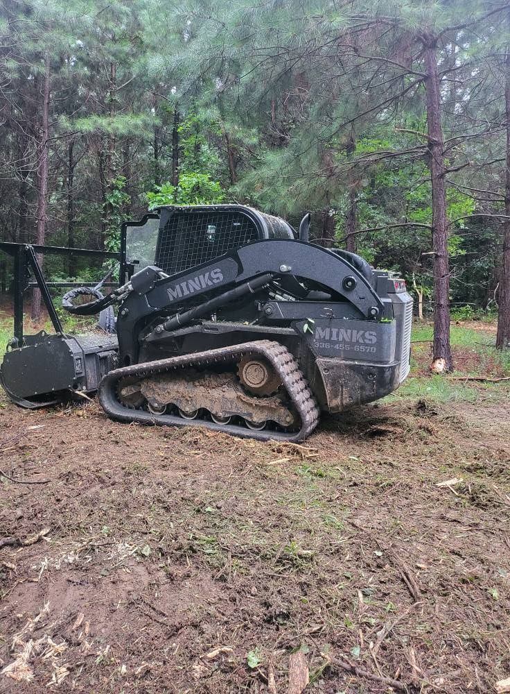 Dark gray tracked skid steer with a forestry mulcher attachment in a forest clearing.