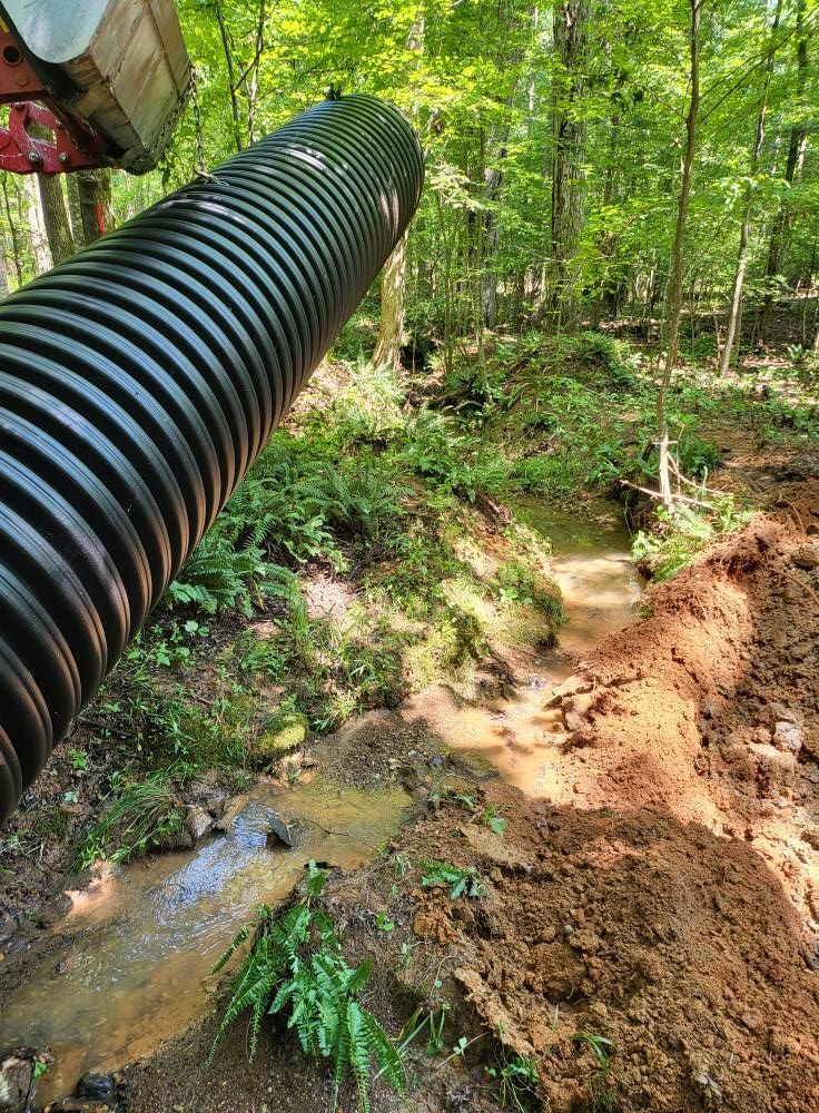 Black corrugated pipe extends over a muddy streambed in a wooded area.