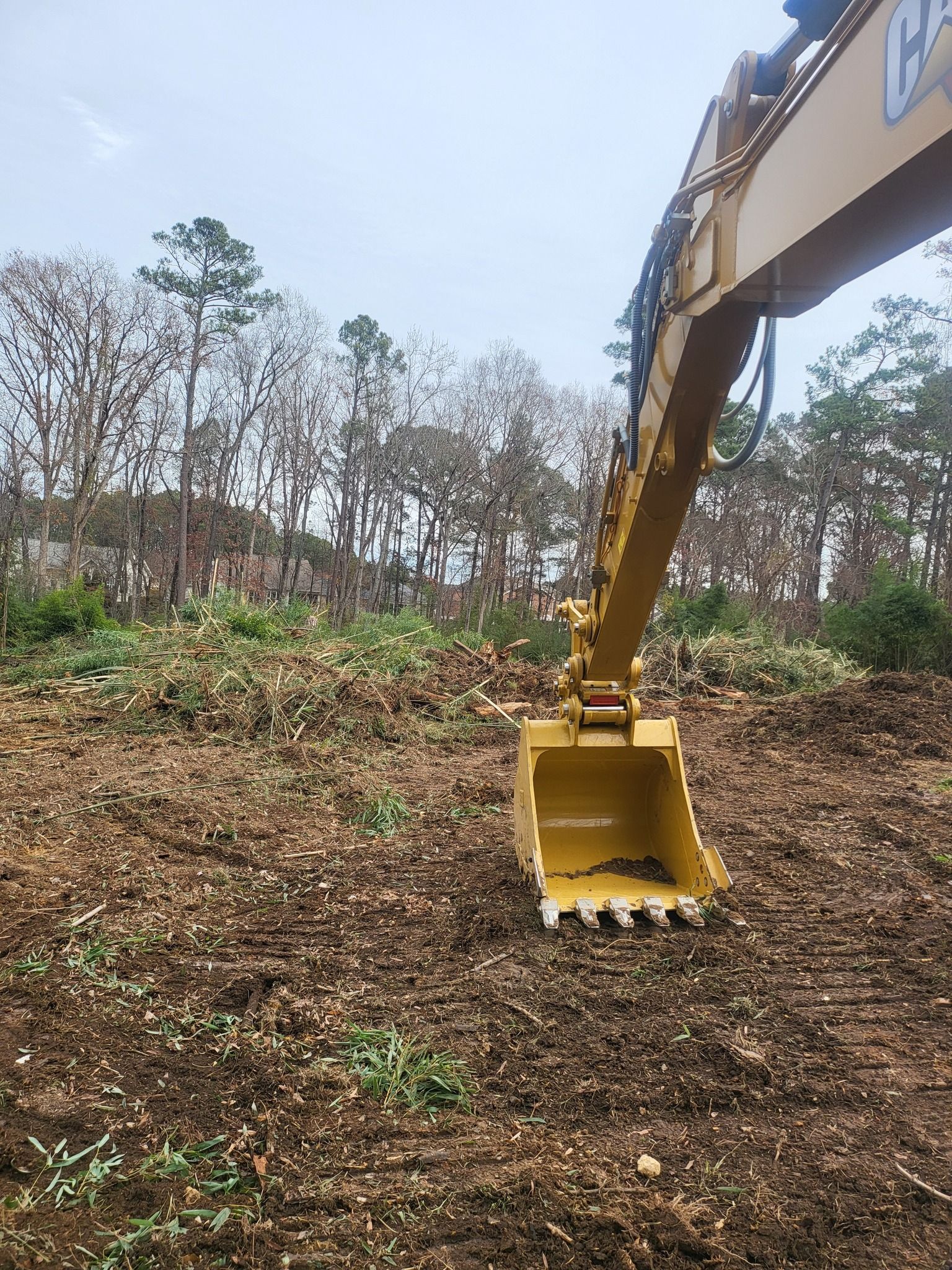 Yellow excavator bucket resting on brown soil in a clearing with trees.