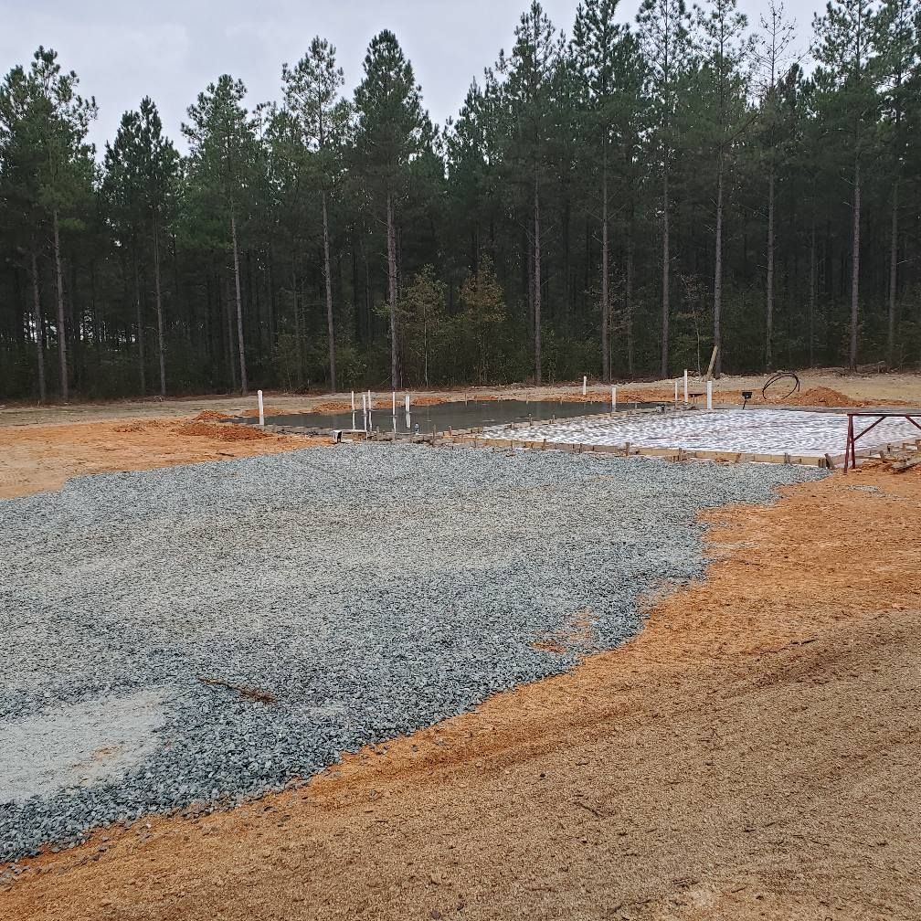 Gravel foundation prepared at a construction site, with exposed pipes and a backdrop of pine trees.