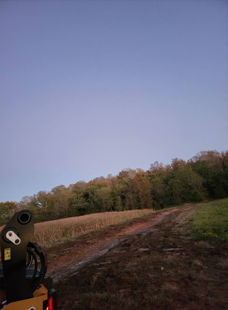 Muddy path leads to a treeline under a pale blue sky. A piece of yellow machinery is in the foreground.