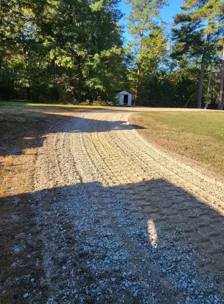 Gravel driveway leading to a small shed surrounded by grass and trees under a clear, sunny sky.