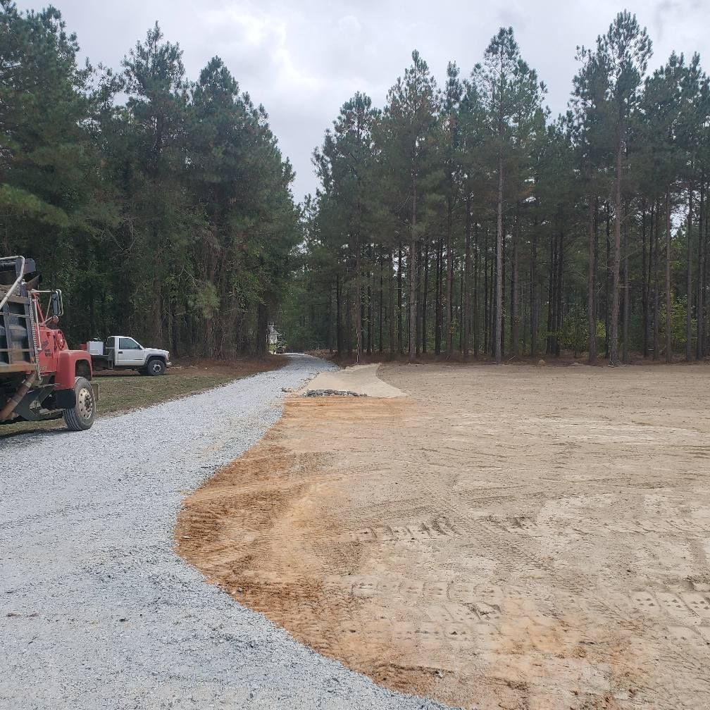 Gravel driveway leading to cleared, sandy area next to a treeline. A truck and tractor are parked on the left.