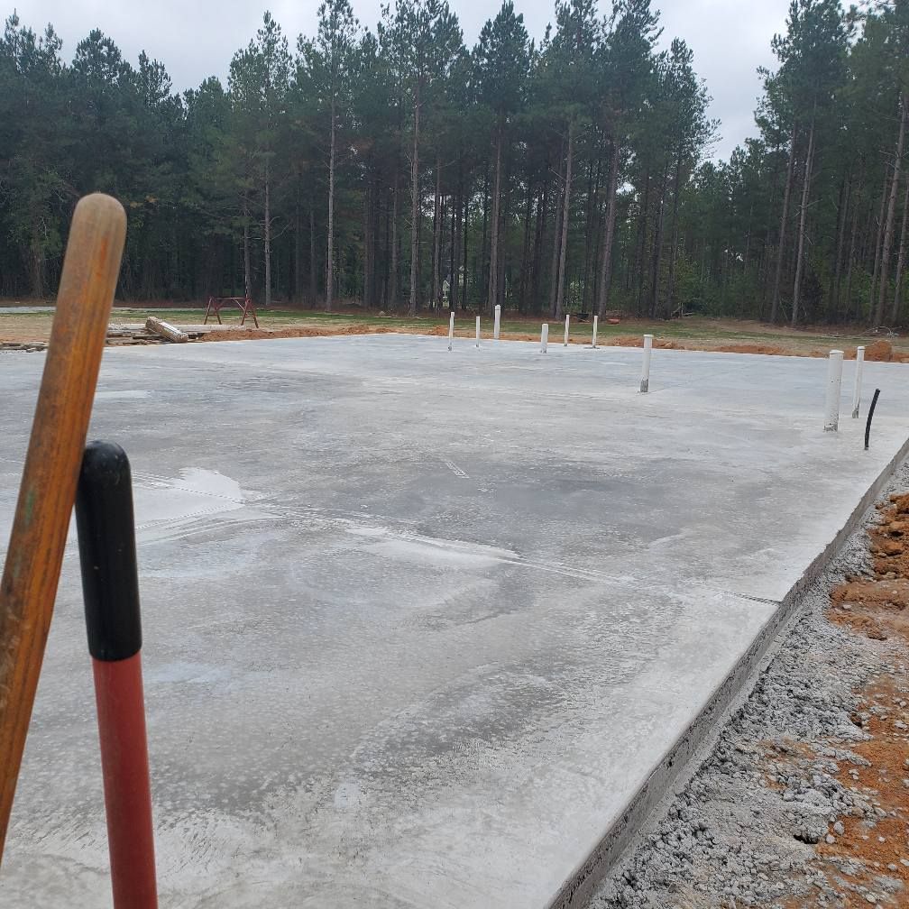 Concrete foundation of a building under construction, with plumbing pipes. Tools lean against it, trees in the background.
