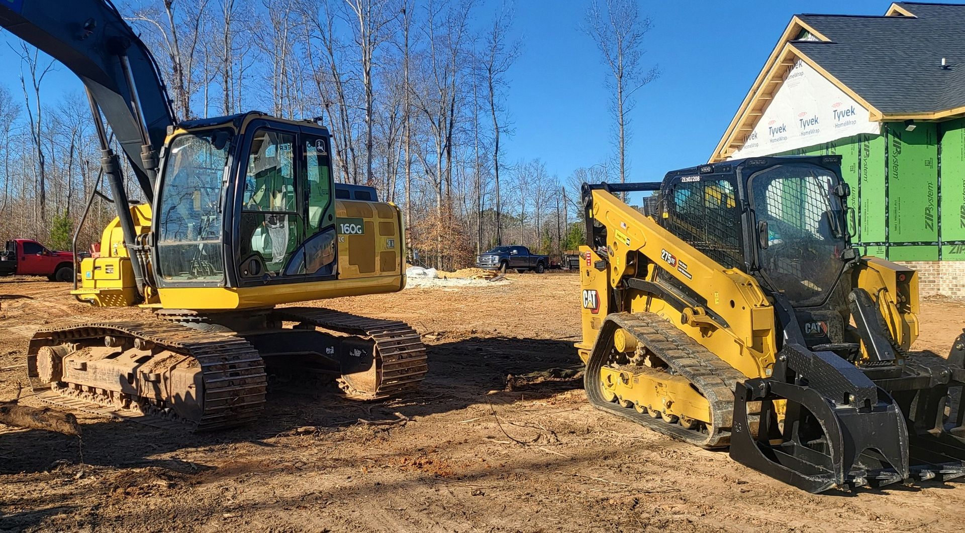 A yellow excavator and a skid steer on a construction site with a partially built house in the background.