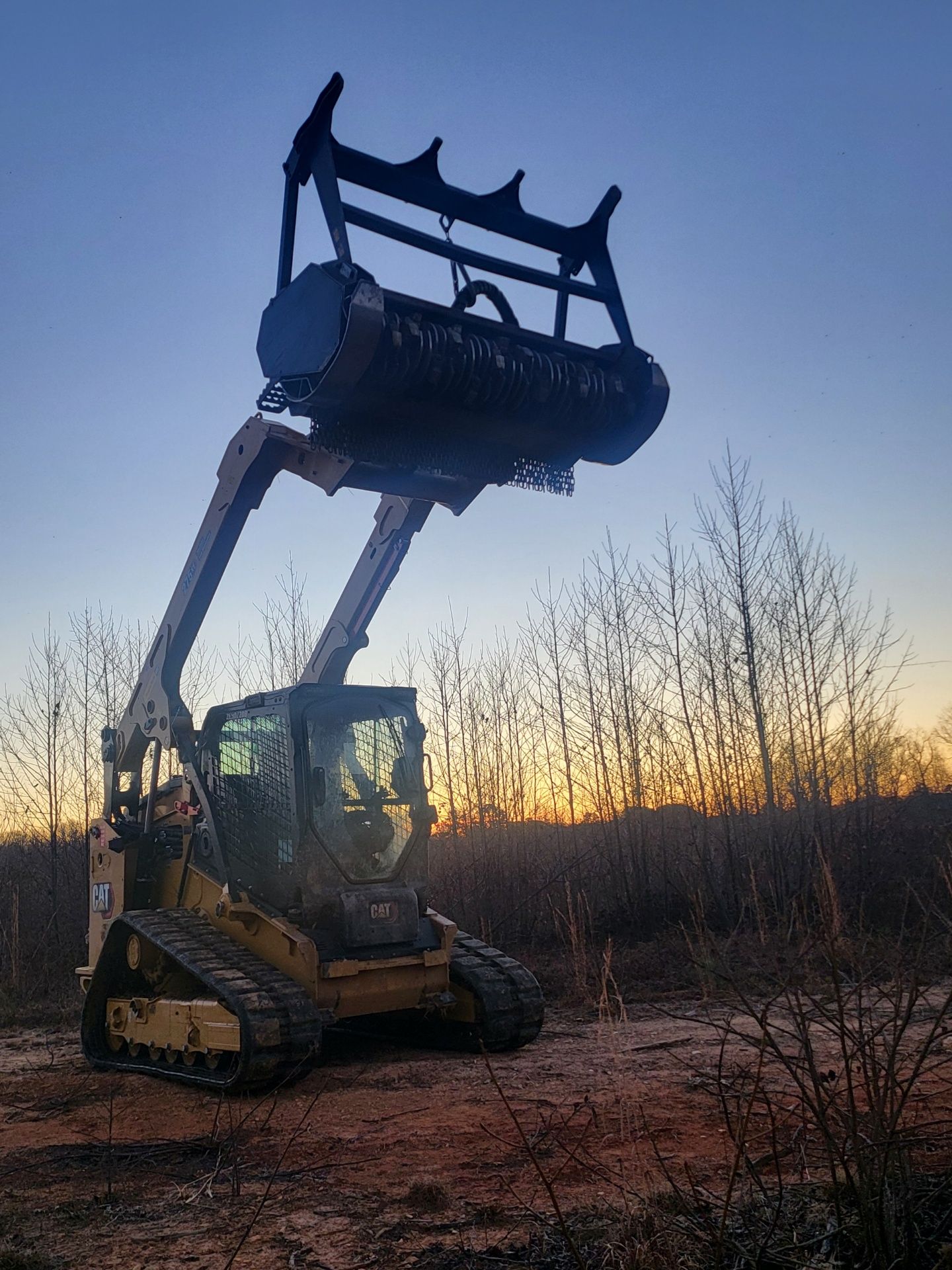 A track loader with a tree shear raised against a sunset.
