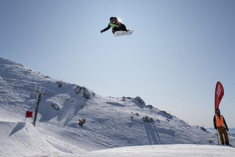 Snowboarder in mid-air above a snow-covered mountain, bright sun, a person with an orange vest observes.
