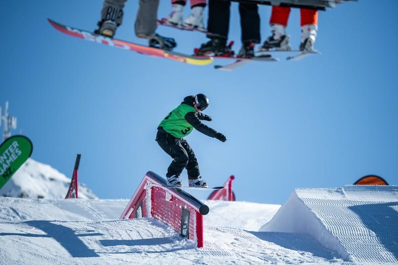 Snowboarder in green and black slides on a rail in a snow park, people on a ski lift overhead.